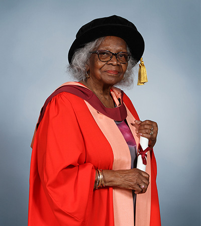 An image of Barbara Dettering wearing a red Honorary Degree gown and holding her degree scroll